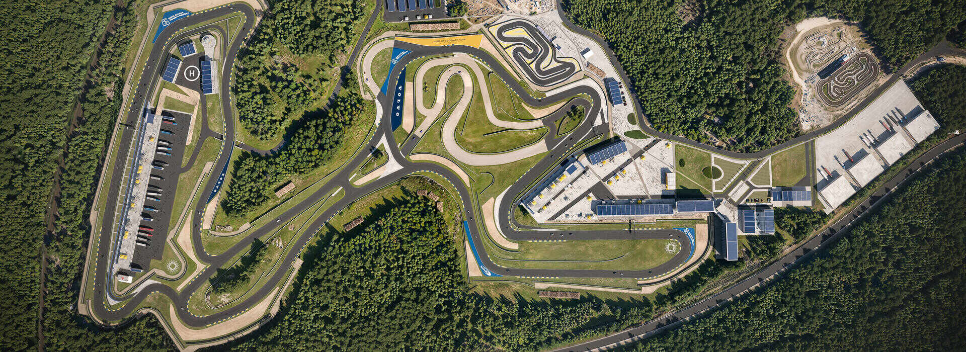 Aerial rendering of a complex motorsport facility surrounded by forest, featuring multiple track layouts, pit buildings, and a helipad — created by omegarender for Apex Circuit Design.